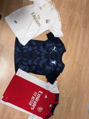 Tre fotbollströjor: Real Madrid, Malmö FF, Arsenal - Säljer tre snygga fotbollströjor: en vit Real Madrid-tröja från Adidas med guldiga detaljer, en blåmönstrad Malmö FF-tröja från Puma och en rödvit Arsenal-tröja från Adidas. Perfekta för fotbollsfans som vill visa sitt stöd på matchen eller träningen.