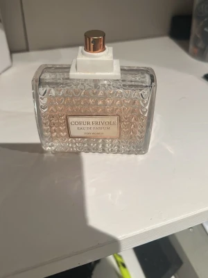 Coeur Frivole Eau de Parfum - Elegant parfymflaska i genomskinligt glas med hjärtformade detaljer och roséguldfärgad spraydel. Innehållet är en ljusrosa Eau de Parfum för kvinnor. Flaskan har en modern, rektangulär form och etikett framtill. Tyvärr inget lock till då det är borttappat..