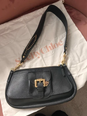 Svart axelremsväska från See By Chloé - Snygg svart axelremsväska från See By Chloé i äkta läder med grov struktur. Väskan har bred axelrem, guldfärgade metalldetaljer och ett dekorativt spänne framtill med See By Chloé-hänge. Nypris över 4000kr