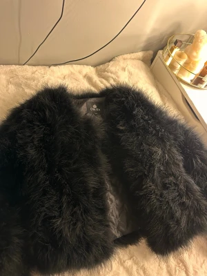 Svart fuskpälsjacka från Chelsea - Säljer en svart, fluffig fuskpälsjacka från Chelsea. Jackan har en kortare modell och är helfodrad med quiltat innerfoder för extra värme. Perfekt för dig som vill ha en trendig och mysig look under vintern. Stängs framtill och har en klassisk, rundad krage.