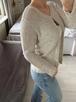 Vit kofta med paljetter - Säljer en vit stickad kofta i storlek M, med små paljettdetaljer och diskreta färginslag. Koftan har rund halsringning, långa ärmar och knäpps framtill med vita knappar. Hör av dig om du undrar något!