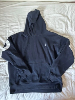 Mörkblå hoodie från Polo Ralph Lauren - Snygg mörkblå hoodie från Polo Ralph Lauren med stor vit logga på ryggen och ärmen. Klassisk huva med knappdetalj och känguruficka framtill. Liten broderad logga på bröstet. Tillverkad i mjukt bomullsmaterial, perfekt för en avslappnad streetwear-look.