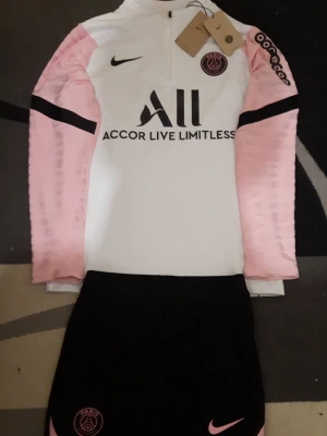 PSG träningsset Nike rosa/svart - Snyggt PSG träningsset från Nike med långärmad vit tröja och rosa ärmar, samt svarta byxor med rosa detaljer. Tröjan har dragkedja, PSG-logga och sponsortryck. Byxorna har PSG-märke och Nike-logga i rosa. Perfekt för fotbollsträning eller chill.
