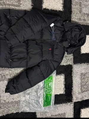 Svart pufferjacka från Polo Ralph Lauren - Svart pufferjacka från Polo Ralph Lauren med röd broderad logga på bröstet. Jackan har huva, flera dragkedjeförsedda fickor och quiltad design. Materialet är polyester och bomull, perfekt för kyliga dagar. Klassisk och stilren look.