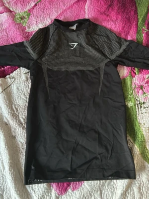 Onyx Gymshark - Säljer en svart träningströja från Gymshark med meshdetaljer upptill och logga på bröstet. Tröjan har en sportig design med inslag av grått och är tillverkad i ett stretchigt syntetmaterial som andas. Perfekt för gymmet eller löprundan.