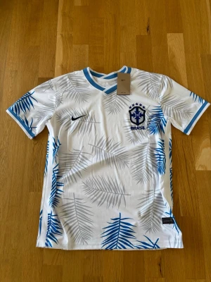Nike Brasil t-shirt med blått mönster - Vit t-shirt från Nike med blå och grå palmbladsmönster, Brasil-logga och blå detaljer vid krage och ärmslut. Tillverkad i ett lätt och ventilerande material, perfekt för sport och träning. Är aldrig använd och är skön att ha på sig. Skicka gärna vid frågor.