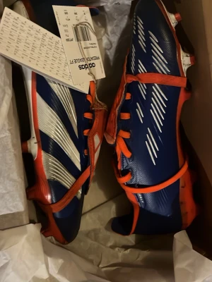 Adidas Predator League FT fotbollsskor - Säljer ett par Adidas Predator League FT fotbollsskor i blått med röda och vita detaljer. Skorna har klassisk snörning, låg profil och mönstrad ovandel för extra grepp på planen. Perfekta för dig som vill sticka ut med både stil och prestanda. Köpte för 600 kr dem är äkta och nya men lite smutsiga. Pris kan deskuteras. 