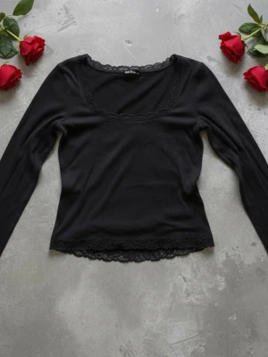Svart långärmad topp med spets - Snygg svart långärmad topp med spetsdetaljer vid halsringning och nederkant. Toppen har en feminin, något tight passform och en klassisk rundad urringning. Perfekt att styla med jeans eller kjol för en trendig look.