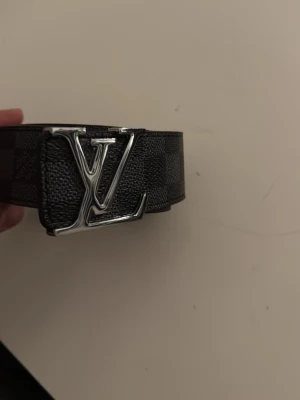 Louis Vuitton rutigt svart bälte helt ny - Snyggt svart bälte från Louis Vuitton med klassiskt rutigt mönster och en stor silverfärgad LV-logga som spänne. Bältet är gjort i skinn och har en exklusiv känsla som lyfter vilken outfit som helst.
