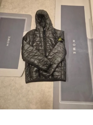 Svart pufferjacka från Stone Island - Snygg svart pufferjacka från Stone Island med huva och dragkedja framtill. Jackan har det klassiska Stone Island-märket på ärmen. Kontakta vid frågor pris går att diskuteras 