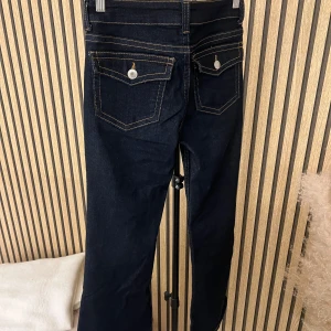 Mörkblå bootcut jeans med ficklock - Snygga mörkblå jeans med bootcut passform och kontrastsömmar. Baksidan har två fickor med lock och knappdetaljer. Klassisk femficksmodell framtill. Jeansen är tillverkade i ett kraftigare denimtyg som ger en tidlös look.