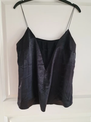 Svart linne med smala axelband - Snyggt svart linne i glansigt tunt material med djup V-ringning och tunna axelband. Perfekt för att styla lager på lager eller bära som det är för en trendig look.