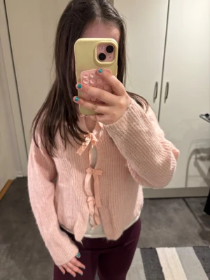 Ljusrosa kofta med rosetter - Supersöt ljusrosa kofta med tre dekorativa rosetter framtill. Koftan är stickad i ett mjukt material och har långa ärmar samt en avslappnad passform. Perfekt för dig som gillar gulliga detaljer och en feminin stil. 