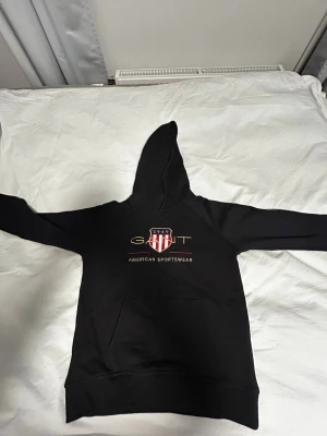 Svart hoodie från GANT  - Svart hoodie från GANT med klassiskt tryck och logga på bröstet i rött, vitt och guld. Hoodien har huva och känguruficka framtill. Mjuk bomullsblandning som är skön att ha på sig. Perfekt för en avslappnad och sportig stil.