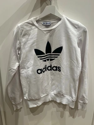 Vit Adidas sweatshirt med logga - Snygg vit sweatshirt från Adidas med den klassiska svarta Trefoil-loggan och texten på bröstet. Tröjan har rund halsringning, långa ärmar och ribbade muddar vid ärmslut och nederkant. Perfekt för en sportig och avslappnad stil.