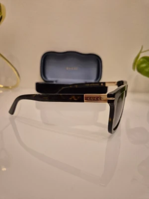 Gucci solglasögon brunmelerad - Solglasögon från Gucci i modellen GG1502S 002. Unisex modell, färg havanna sköldpaddsfärgad båge med logga i guld på sidorna. Bågbredd 143 mm. Oanvända, nya! Köpta på Mister Spex i höstas, pris på hemsida: 3899 kr Kvitto ses på sista bilden. Kommer i orginalfodral i svart sammet med putsduk mm.