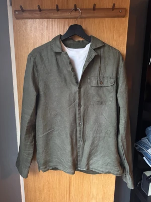 Linne skjorta från Riley  - Snygg olivgrön skjorta i linne med klassisk krage och knappstängning framtill. Skjortan har en bröstficka med knapp och långa ärmar med manschett. Perfekt för en avslappnad men stilren look.