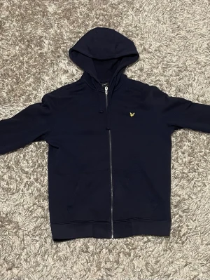 Mörkblå hoodie från Lyle & Scott - ( kan passa M )Snygg mörkblå hoodie med dragkedja från Lyle & Scott. Klassisk design med huva och två fickor framtill. Liten gul logga på bröstet ger en clean look. Tillverkad i mjukt bomullsmaterial som känns skönt mot huden. Perfekt för en avslappnad stil.