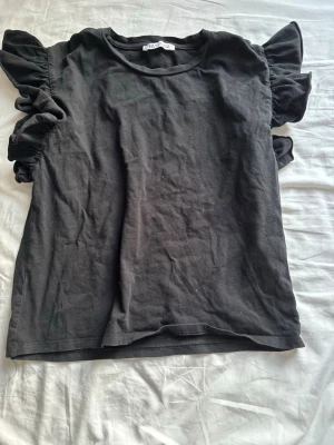 Svart topp med volangärmar från Zara - Säljer en svart topp från Zara med volangdetaljer på ärmarna. Toppen har rund halsringning.