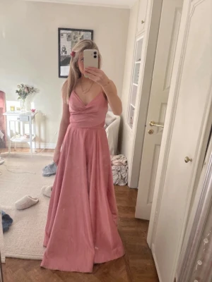 Lång rosa klänning med v-ringning - Elegant lång klänning i rosa med v-ringad omlott överdel och breda axelband. Klänningen har markerad midja och ett flödande, utsvängt kjolparti. Perfekt för student eller bal. Materialet ser ut att vara satin eller polyester. Tyvärr lite smutsig där nere, passar om man är cirka 165
