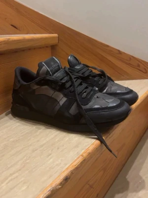 Valentino rockrunners - Snygga svarta sneakers från Valentino med camo-mönster i olika nyanser av svart och grått. Skorna har låga skaft, svarta skosnören och detaljer i både läder och textil. Perfekta för dig som vill ha en stilren men ändå unik look köpte för 2400 kr 11 månader sen storlek 42 men sitter som 41