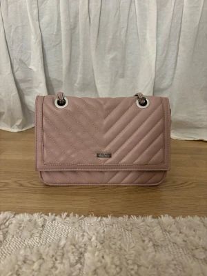 Rosa quiltad handväska med kedjerem - Jättefin handväska från Don donna! Den är ljusrosa och har många bra praktiska egenskaper, 3 st fack varav 2 med dragkedja. Man kan även välja om man vill ha långt eller kort band. Den stängs med magneter och i helhet en väldigt praktisk väska! Fin skick förutom en liten missfärgning på baksidan men inget som syns om man inte kollar noga. 