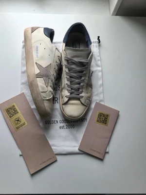 Golden Goose Superstar vita sneakers - Golden Goose Superstar sneakers i vitt läder med grå mockadetaljer och blå hälflik. Klassisk stjärndesign på sidan och perforerad tå. Skorna har grå skosnören och slitna detaljer för en trendig look. Kommer med originalpåse och certifikat.