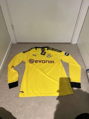 Gul långärmad BVB Puma tröja - Säljer en gul långärmad Borussia Dortmund-tröja från Puma med svarta detaljer och klubbmärke på bröstet. Tröjan har trycket 'Evonik' på framsidan och 'Dortmund' på ryggen. Perfekt för fotbollsfans som vill visa sitt stöd på matchen eller träningen.