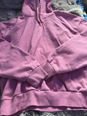 Rosa hoodie med huva och dragsko - En klassisk rosa hoodie med huva och dragsko. Hoodien har ribbade muddar vid ärmslut och nederkant samt en avslappnad passform. Perfekt för en chill och avslappnad stil.