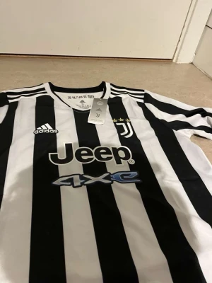 Juventus långärmad matchtröja Adidas - Juventus långärmad matchtröja i svartvitt med klassiska ränder, Adidas-logga och Jeep-sponsring på bröstet. Tröjan har rund hals, tre stjärnor ovanför klubbmärket och är tillverkad i ett lätt, sportigt material perfekt för fotboll.