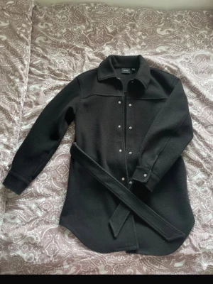 Svart overshirt med bälte från Gina Tricot - Stilren svart overshirt från Gina Tricot med knappar framtill och ett matchande bälte i midjan. Overshirten har klassisk krage, långa ärmar och är gjord i ett mjukt, ull-liknande material. Perfekt för lager på lager och enkel att styla.