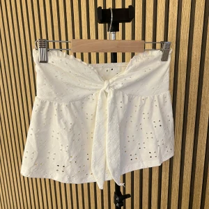 Vit topp med knyt och broderad spets - Superfin vit topp i bomull med broderad spets och hålmönster. Toppen har en söt knytning framtill och volangkant som ger en luftig och somrig känsla. Perfekt för varma dagar och lätta outfits.