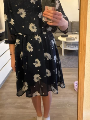 Mörkblå blommig kortklänning - Söt mörkblå kortklänning med vita blommor och transparenta ärmar. Klänningen har resår i midjan, rund halsringning och en öppen detalj i ryggen. Perfekt för dig som gillar en luftig och feminin stil.