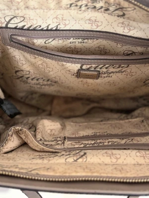 Vit handväska från Guess - Snygg vit/grå handväska från Guess med långa bruna axelband och silverdetaljer. Väskan har ett rymligt huvudfack, flera innerfickor och Guess-logga både på utsidan och insidan. Tillverkad i konstläder med mönstrat tygfoder.