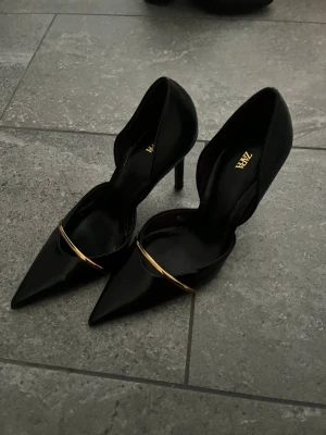 Svarta pumps med gulddetalj från Zara - Snygga svarta pumps från Zara med spetsig tå och elegant guldfärgad detalj över foten. Skorna har hög klack och öppen insida för en modern look. Tillverkade i glansigt material som ger en lyxig känsla.