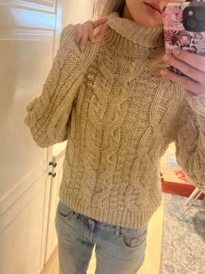 Beige kabelstickad polotröja - Säljer en chunky beige kabelstickad polotröja med bred krage och långa ärmar. Tröjan har ett klassiskt flätmönster och är gjord i ett mjukt, tjockt garn som håller dig varm. Perfekt för kyliga dagar och enkel att styla med jeans.