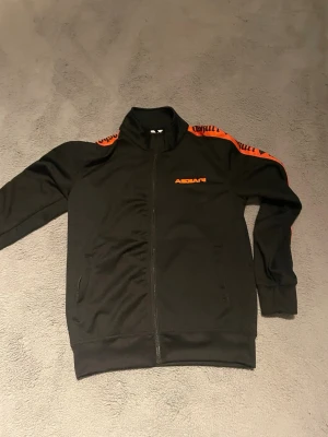 Svart track jacket från Ascari - Svart track jacket från Ascari med dragkedja framtill och orange logga på bröstet. Snygga orange detaljer och text längs axlar och ärmar. Tröjan har ståkrage och ribbade muddar vid ärmslut och nederkant. Perfekt för en sportig och trendig look.