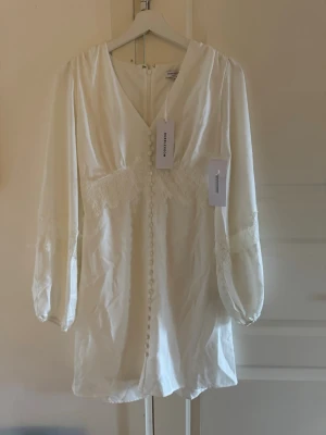 Vit spetsklänning, perfekt till studenten  - Supersöt vit kortklänning från Bubbleroom med v-ringning, spetsdetaljer över byst och ärmar samt dekorativa knappar framtill. Klänningen har långa ärmar med lätt puff och är gjord i ett mjukt, glansigt material som känns lyxigt.