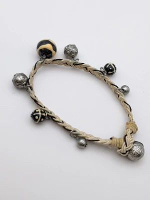 Flätat armband med pärlor och berlocker - Unikt armband med flätad beige och svart tråd, dekorerat med flera pärlor i silverfärg samt svarta och beige mönstrade pärlor. Armbandet har små berlocker som ger en cool och personlig touch. Perfekt accessoar för dig som gillar detaljer.