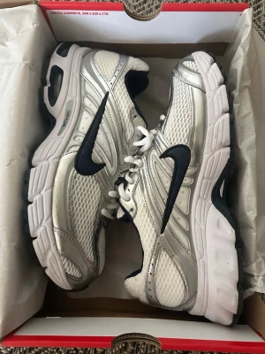 Nike silvervita sneakers med svart swoosh - Nike sneakers i silver och vitt med svart swoosh-logga på sidorna. Ovandelen är i mesh och syntetmaterial för bra andningsförmåga och komfort. Tjock, vit sula med svarta detaljer och klassisk snörning. Perfekta för en sportig och trendig look. Helt nya använda bara en gång paket ingår