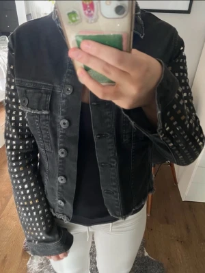 Svart jeansjacka med nitar - Cool svart jeansjacka med silvriga fyrkantiga nitar längs hela ärmarna. Klassisk knappstängning framtill och två bröstfickor med lock. Jackan har en rå och edgy känsla, perfekt för att sticka ut i mängden.