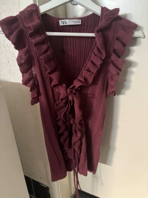 Vinröd ribbad topp med volang från Zara - Supersnygg vinröd ribbad topp från Zara med volangdetaljer längs ärmhålen och v-ringningen. Toppen har snörning framtill och är ärmlös, perfekt för att lyfta vilken outfit som helst. Är storlek M men passar toppen som S