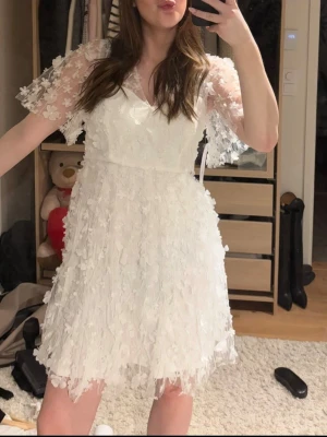 Studentklänning - Supersöt vit kortklänning med 3D-blommor och volangdetaljer. Klänningen har genomskinliga korta ärmar med blommönster och v-ringad hals. Perfekt för sommarens alla tillfällen och studenten! ☀️❤️ nypris 999kronor. 