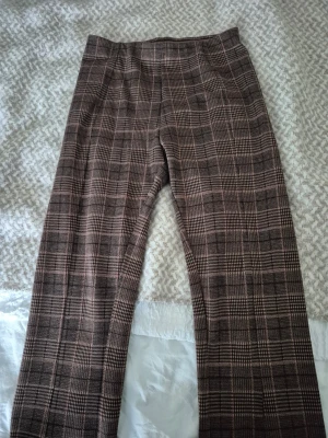 Bruna rutiga kostymbyxor - Snygga bruna kostymbyxor med klassiskt rutigt mönster i beige och mörkbrunt. Byxorna har rak passform och hög midja, perfekta för en stilren look. Materialet känns mjukt och har en lätt struktur. Inga synliga fickor eller detaljer.