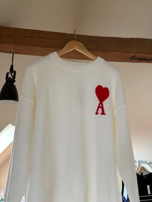 Vit stickad tröja från Ami Paris - Vit stickad tröja från Ami Paris med rund halsringning och ribbade muddar. På bröstet finns Ami:s ikoniska röda hjärta med bokstaven 'A'. Tröjan har en avslappnad passform och är tillverkad i mjukt material, perfekt för en stilren look.