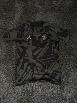 Svart PSG  träningströja - Säljer en svart och grå PSG träningströja i samarbete med Jordan. Tröjan har ett grafiskt mönster, rund hals och korta ärmar. PSG-logga och Jumpman-logga på bröstet. 