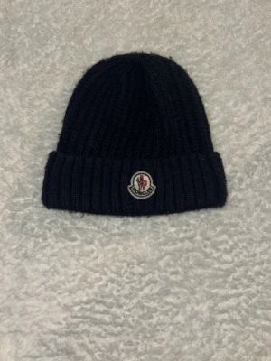 Svart ribbstickad mössa från Moncler - Säljer en svart ribbstickad mössa från Moncler med uppvikt kant och klassisk Moncler-logga framtill. Mössan är gjord i mjukt material och har en stilren, enkel design som passar till det mesta. Pris diskuteras!