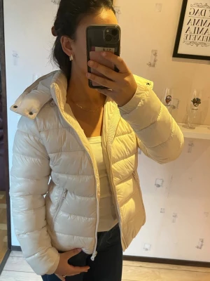 Vit pufferjacka med hög krage - Säljer en snygg vit pufferjacka med hög krage och dragkedja. Jackan har quiltad design, två sidofickor med dragkedja och en avtagbar huva med tryckknappar. Perfekt för våren. 