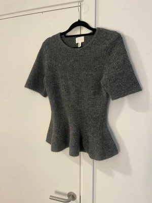 Grå stickad topp med volangkant från H&M - Snygg grå stickad topp med korta ärmar och volangkant nedtill. Tröjan har rund halsringning och är ribbstickad för en skön och stretchig passform. Perfekt till soliga vårdagar!