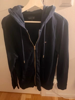 Mörkblå zip hoodie från Lexington - Mörkblå hoodie från Lexington med dragkedja framtill och justerbar huva med snören. Klassisk design med fickor på sidorna och diskret Lexington-logga på bröstet. Tillverkad i mjukt bomullsmaterial som känns skönt mot huden. Den är använd ett fåtal gånger bara